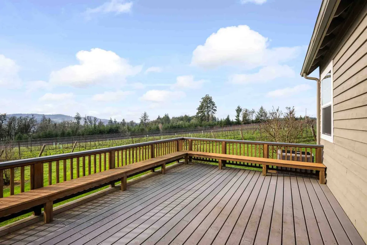 A spacious wooden deck with comfortable seating, set against a sweeping landscape of vineyards and distant mountains. The area is arranged for relaxation and gatherings, with ample space for enjoying the outdoors, and the sun sets behind the mountains, casting a warm glow over the scene.