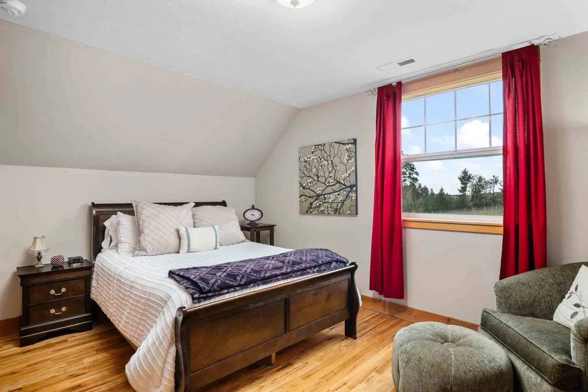 This image presents a welcoming bedroom with a bed, window, tasteful artwork, and a chair. The room is bathed in gentle light, inviting relaxation and quiet moments, while the window hints at scenic countryside vistas.