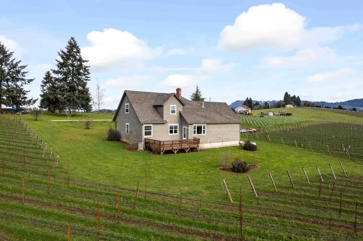 A wide exterior view of the house set against a backdrop of vineyards and surrounding trees, with a spacious deck perfect for gatherings. The mood is serene and inviting, ideal for relaxation and family-oriented activities like outdoor meals or watching sunsets.