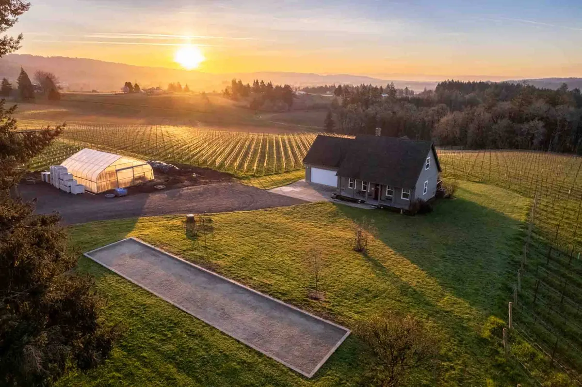 The image showcases a charming single-family farmhouse set against the sweeping backdrop of rolling vineyards and golden sunset skies. The home’s inviting porch overlooks a manicured bocce court, with lush greenery and rows of grapevines stretching into the distance. The atmosphere is tranquil and welcoming, hinting at relaxing evenings, wine country adventures, and leisurely outdoor activities.