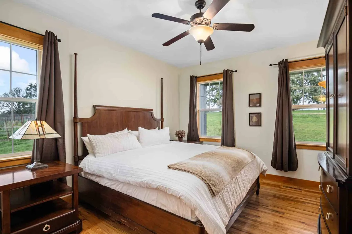 This image features a comfortable bed, a nightstand, windows allowing in morning light, and a ceiling fan. The scene exudes a sense of calm and relaxation, with natural light streaming through the windows and soft bedding inviting rest.