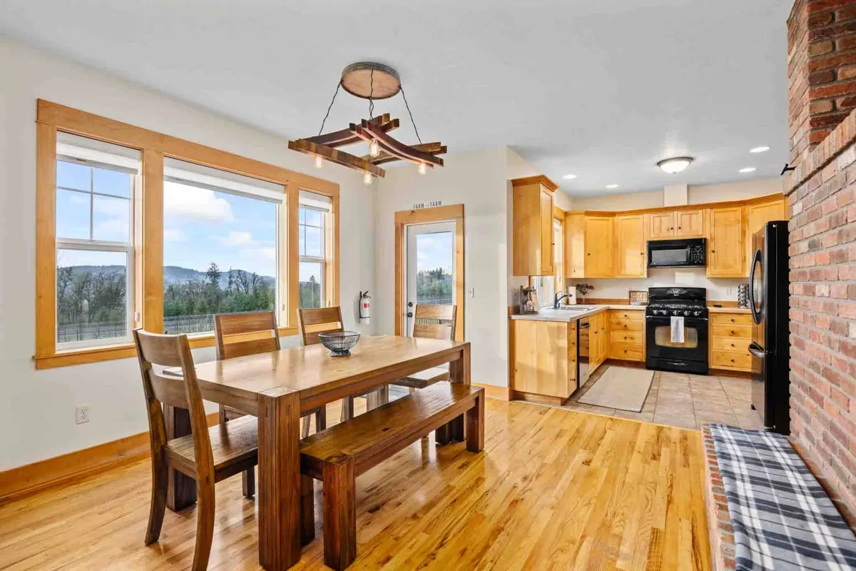 A warm, inviting shot focused on the dining table set against large windows and the connected kitchen area. The hardwood floors, brick wall, and light streaming in create a cozy atmosphere for family meals or holiday celebrations, with scenic vineyard vistas just outside.