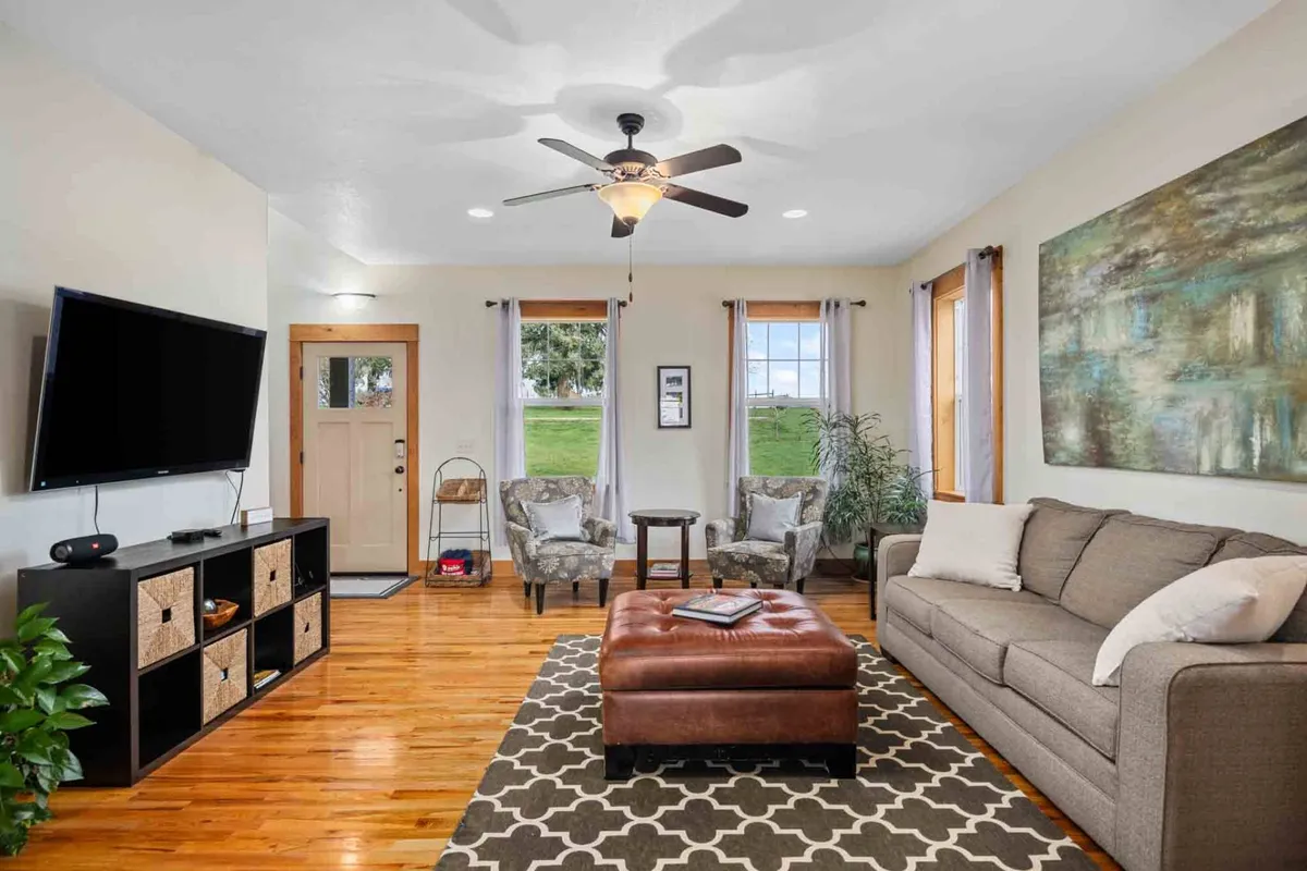 A comfortable living room featuring a sofa, coffee table, TV, artwork, and large windows. The setting exudes a welcoming mood, perfect for relaxing, entertaining guests, or family game nights, with views of the surrounding landscape visible.