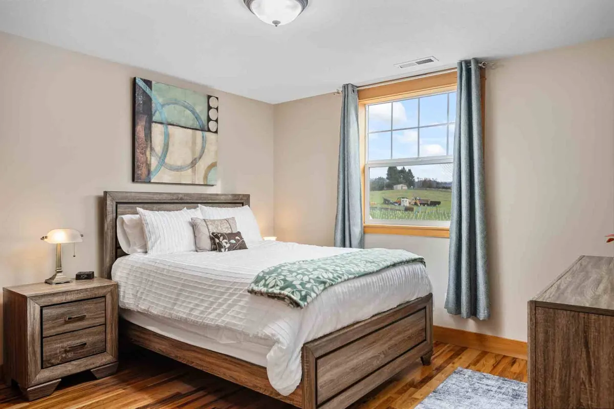 A cozy bedroom is shown with a neatly made bed in white linens, an art piece on the wall, a window with views to the outside, and a nightstand with a lamp. The scene feels peaceful and filled with natural light, offering a sense of quiet retreat.